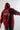 A person viewed from behind wears a deep red hoodie with the hood up against a light gray background. A graphic on the upper back features a white paint-splatter shape, a red-orange silhouette with a circular emblem, and the word "HIMEKO" in prominent white text.