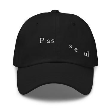 Front view of a baseball cap in black. The words "pas seul" are embroidered across the front panel in white, the letters set in a fine serif and scattered so they step down and to the right. The cap is an unstructured low-profile six-panel in smooth chino twill, with embroidered eyelets and an adjustable metal buckle strap.