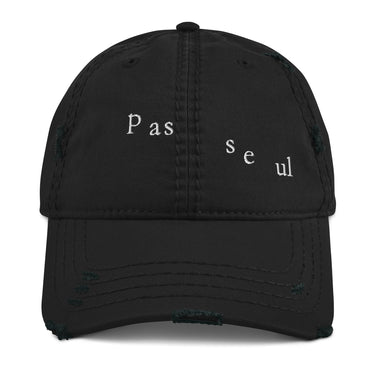 Front view of a distressed hat in black. The words "pas seul" are embroidered across the front panel in white, the letters set in a fine serif and scattered so they step down and to the right. The cap is an unstructured low-profile six-panel with a curved, stitched brim and a soft pre-shrunk twill crown.