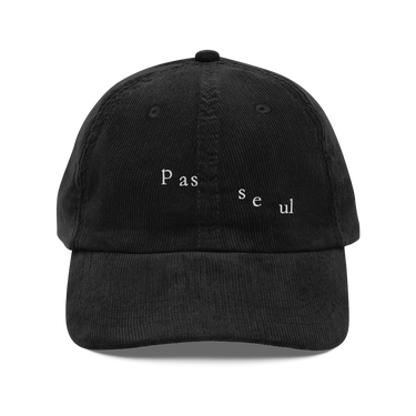 Front view of a corduroy cap in black. The words "pas seul" are embroidered across the front panel in white, the letters set in a fine serif and scattered so they step down and to the right. The cap is an unstructured low-profile six-panel in ridged cotton corduroy, with embroidered eyelets and a metal buckle strap.