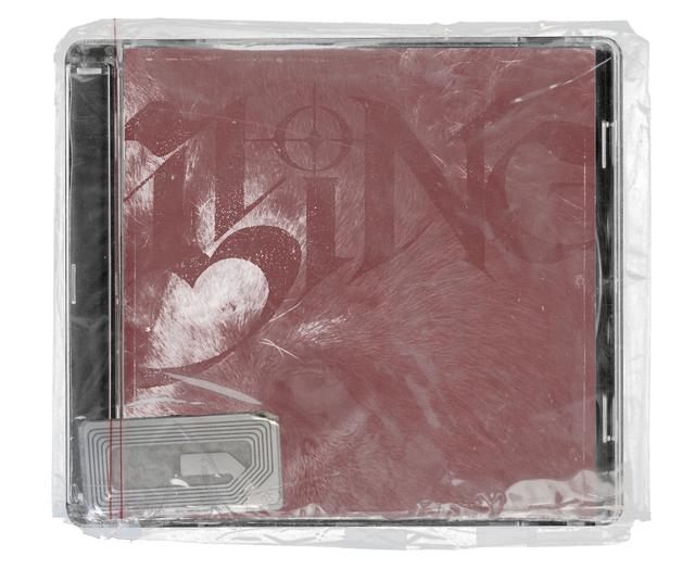 CD jewel case in clear shrink wrap. The cover is a close-up of a lion's fur and eye in a dusty red monotone, overlaid with large decorative lettering and a crosshair motif, with a silver security tag in the lower corner.