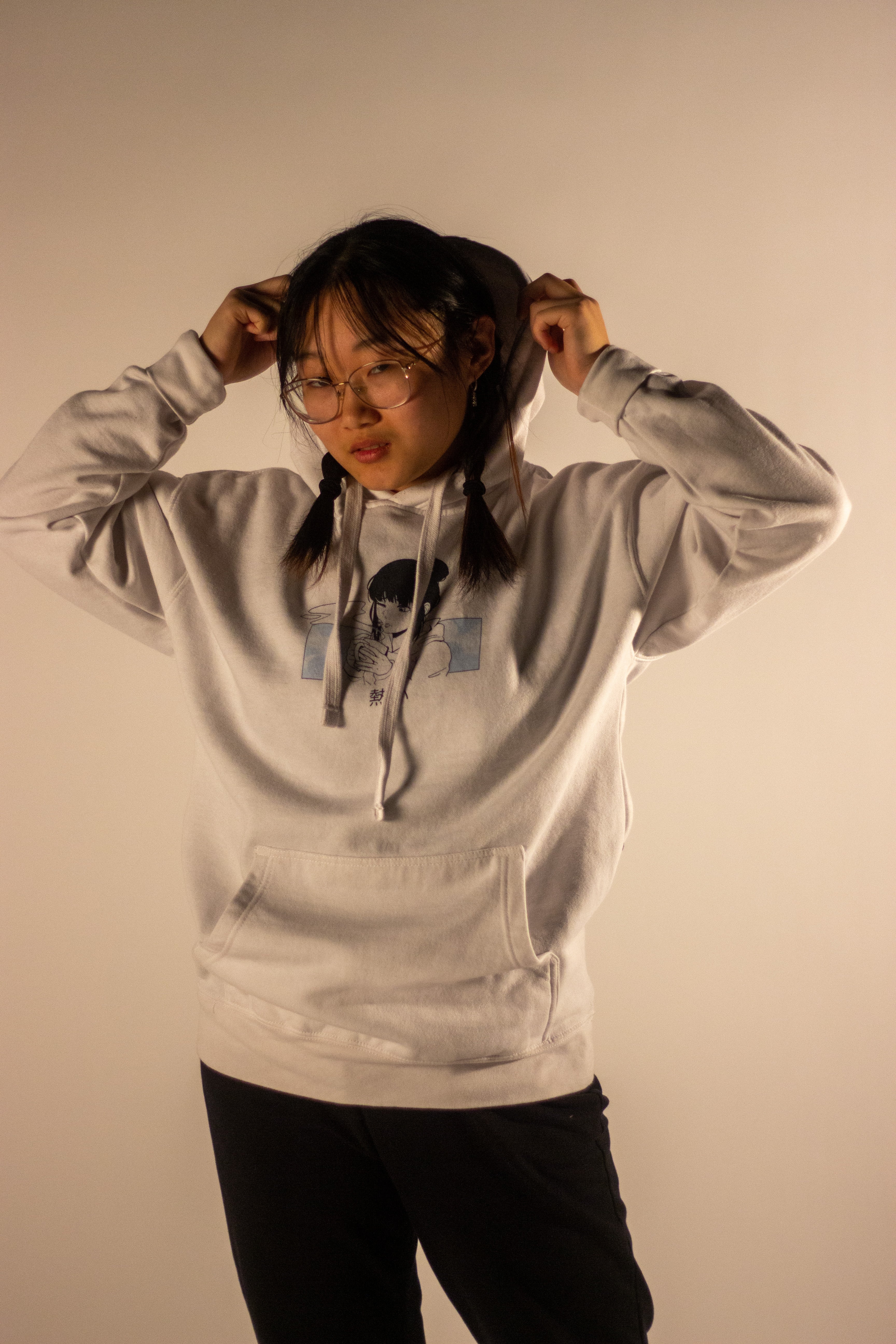 A model with glasses puts on the hood of a light grey hoodie. The hoodie has a small rectangular graphic on the chest depicting an anime character in a light blue, manga-style panel.