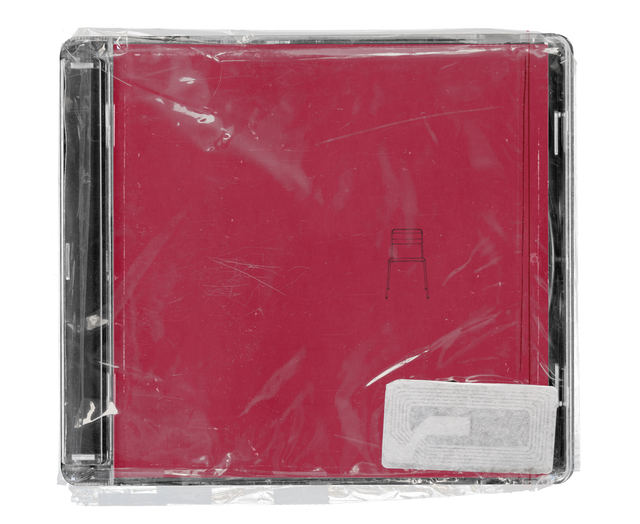 CD jewel case in clear shrink wrap. The cover is plain crimson with a tiny line drawing of a chair at its centre, and a silver security tag in the lower corner.