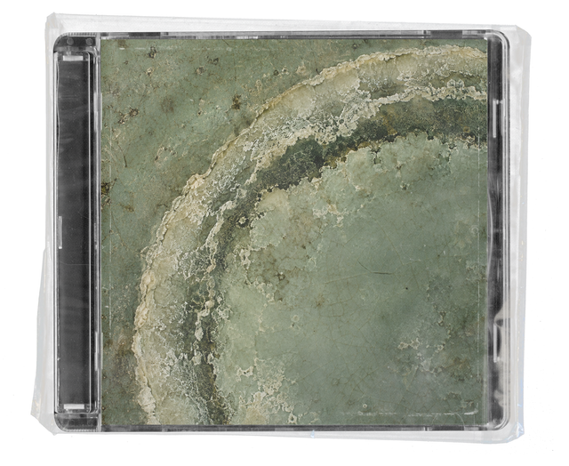 CD jewel case in clear shrink wrap. The cover is a close-up of weathered green stone with a sweeping, pale, crusted arc running across it like a mineral ring.