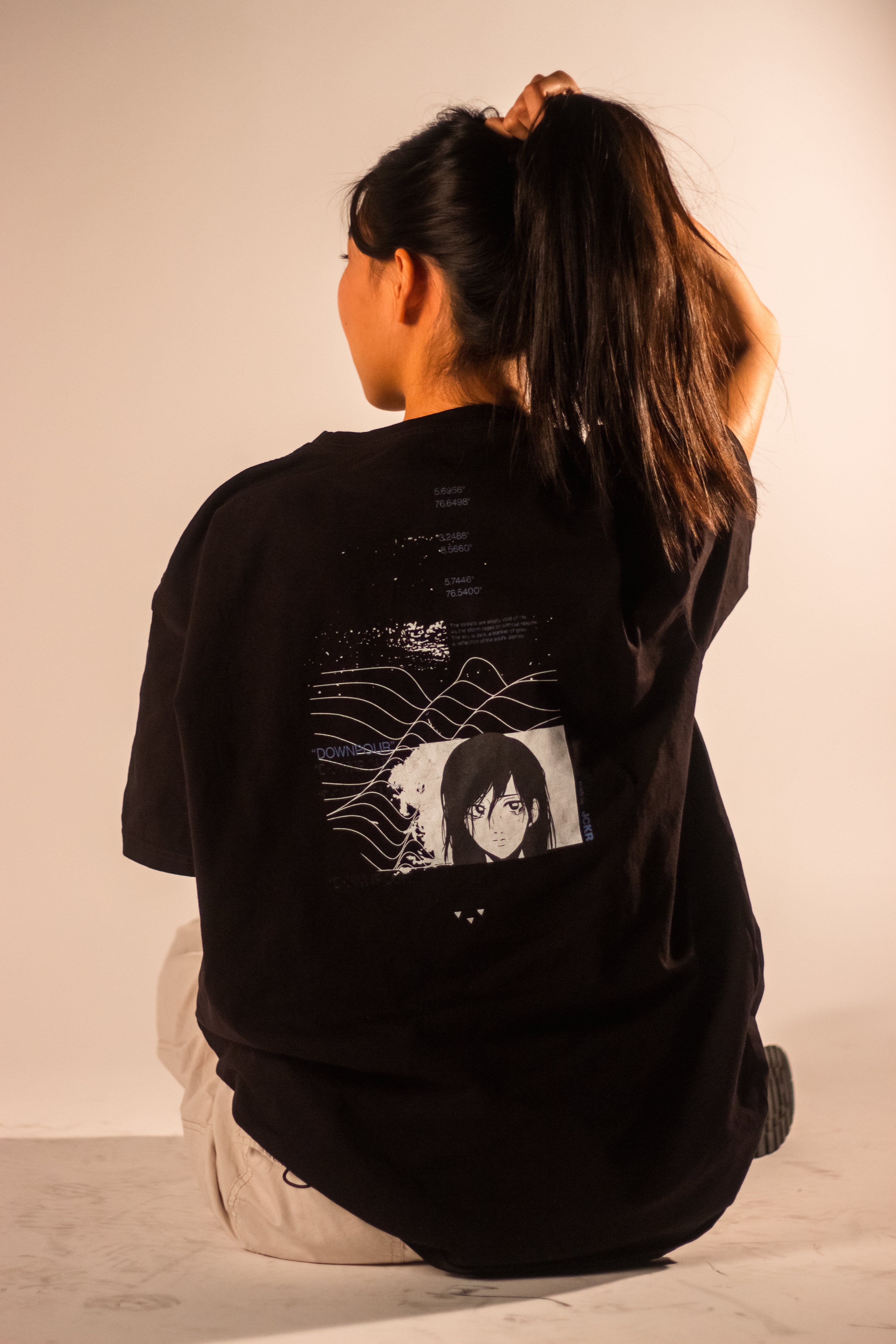 A model, with her back turned, gathers her dark hair to showcase the graphic on a black oversized t-shirt. The design has a tech-inspired look, with a rectangular portrait of an anime character below a field of white, wavy contour lines.