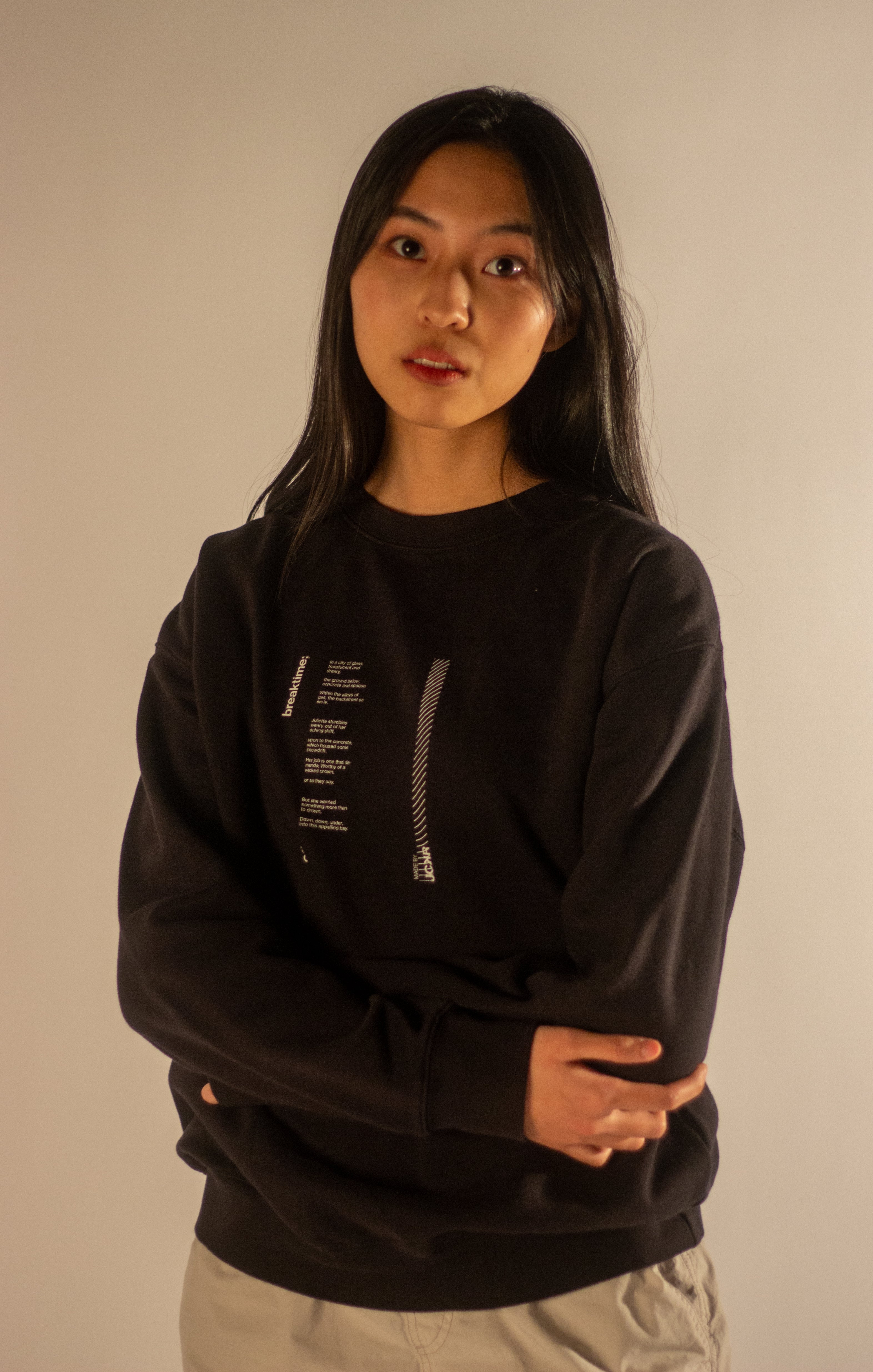 Front view of a model wearing a black crewneck sweatshirt. The shirt has a minimalist graphic on the left chest featuring a vertical column of small white text next to a stylized silver zipper.