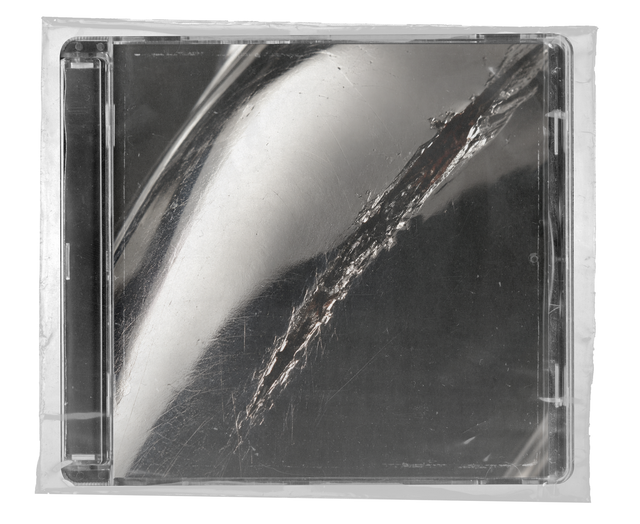CD jewel case in clear shrink wrap. The cover is a close-up black-and-white photograph of polished metal with a bright curved highlight and a jagged scratch running diagonally across it.