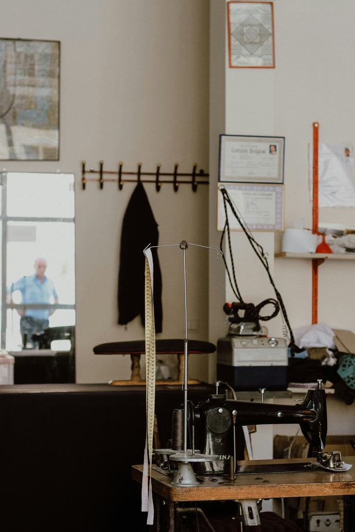 A vertical shot inside a tailor's or seamstress's workshop. In the foreground, an antique-looking black sewing machine sits on a wooden table. A metal stand with a long, yellow measuring tape hanging from it is positioned next to the sewing machine. In the background, a dark jacket hangs on a wall rack. To the left, a mirror reflects a person in a blue shirt. Various items, including framed certificates and possibly other sewing equipment, are visible on the walls and shelves around the room.
