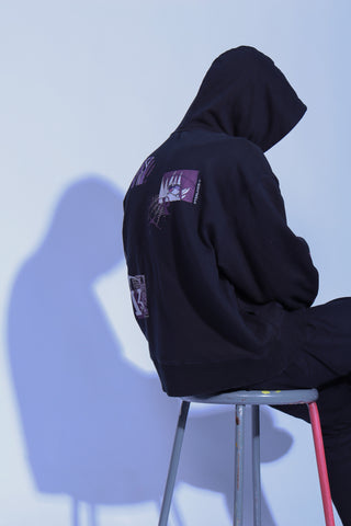 A person sits on a stool with their back to the camera, wearing a solid-colored hoodie with the hood up. The back of the hoodie features several scattered, high-contrast graphics with an abstract, "glitch" aesthetic.