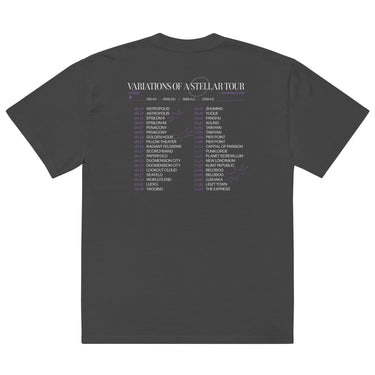 Back view of an oversized tee in faded black. A large graphic fills the upper back, laid out like a concert tour poster. A wide display serif title reads "VARIATIONS OF A STELLAR TOUR" in white, with a small italic "robin" and "/summeretto" tucked beneath it and a ruled row of four era markers below that. Two columns of dated stops run down the rest in condensed capitals, the dates set in violet, with thin outlined starbursts drifting across the list.