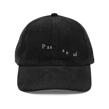 Front view of a corduroy cap in black. The words "pas seul" are embroidered across the front panel in white, the letters set in a fine serif and scattered so they step down and to the right. The cap is an unstructured low-profile six-panel in ridged cotton corduroy, with embroidered eyelets and a metal buckle strap.