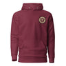 Front view of a solid-colored hoodie. A small, embroidered crest is on the left chest, featuring a central symbol within a circular and star-like frame.